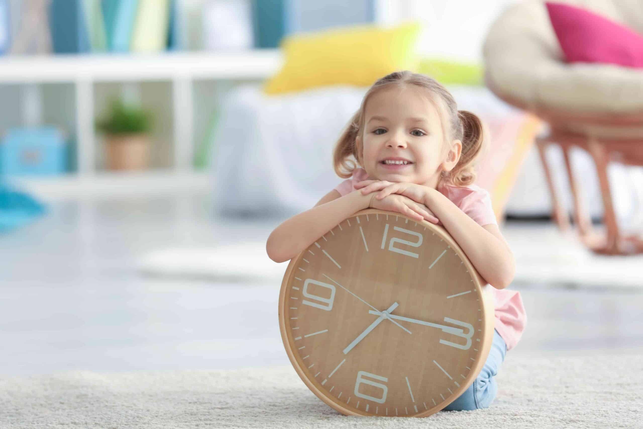 Smiling young girl holding a big clock, while sitting on floor at home. Representative of time with each parent and the topic of parenting plans and parenting agreements for separated parents.